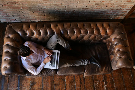 A guy on the sofa of his loft, works or studies at the computer by sending work mails or working on a design project. Concept of: work, technology, design and university student.の写真素材