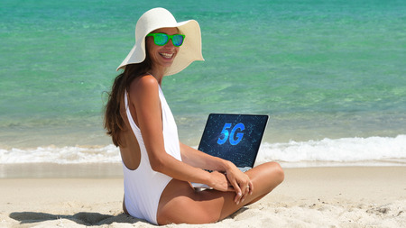 Beautiful woman is sitting on the sand, working at the computer with a white bath, in a white bathing suit.の写真素材