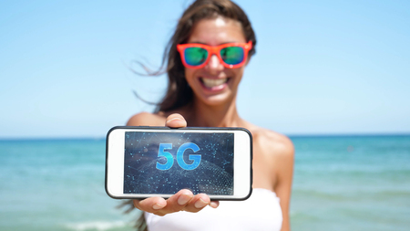 Beautiful girl laughs happily and holds the phone with 5G internet, on the sea in sunglasses, in a white bathing suit.の写真素材