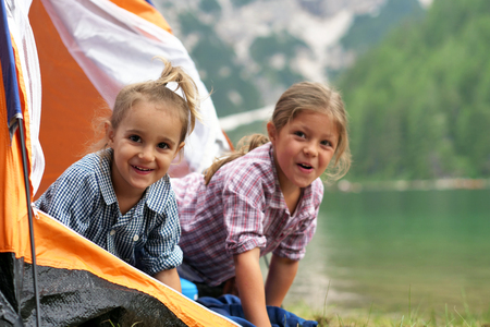 Two sisters camping at the lake, they enjoy playing in the tent, surrounded by nature. Concept of: sisterhood, travel, camping for children.の写真素材