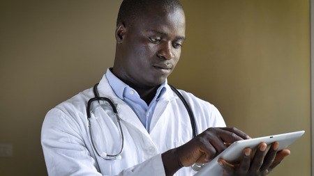 A successful doctor smiles (African American), works in a tablet, wearing a white coat, stethoscope, brown eyes, brown background.の写真素材