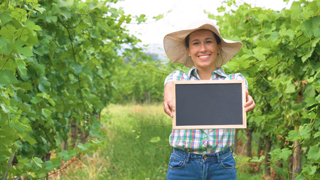 Beautiful girl (woman) farmer smiling watching the grape fields, holding a black board, in a shirt, greens backgroundの写真素材