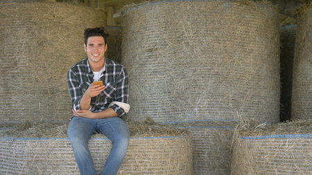 A handsome young farmer (student) is sitting on a haystack, talking on the phone, writing a message, selfie, on a shirt, happy smiling.の写真素材