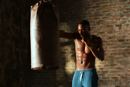 An Afro-american guy trains in a vintage gym and fists his boxing bag fists. Concept of: gym, fitness, boxing, success, workout and powerの写真素材