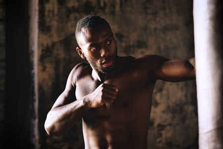 An Afro-american guy trains in a vintage gym and fists his boxing bag fists. Concept of: gym, fitness, boxing, success, workout and powerの写真素材