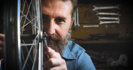 Well-groomed handsome bearded master hipster, specialist in bicycles, repairing a bicycle in his workshop, wheels, frames, spokes, the background of tools. Concept: pro bike, cycle passion, lifestyle.の写真素材