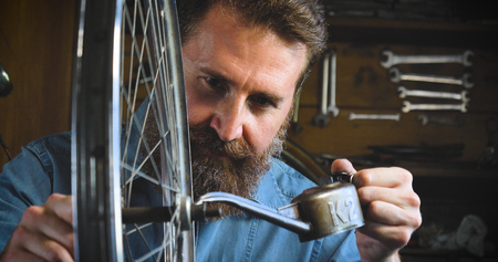 Well-groomed handsome bearded master hipster, specialist in bicycles, repairing a bicycle in his workshop, wheels, frames, spokes, the background of tools. Concept: pro bike, cycle passion, lifestyle.の写真素材