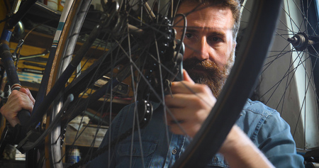Well-groomed handsome bearded master hipster, specialist in bicycles, repairing a bicycle in his workshop, wheels, frames, spokes, the background of tools. Concept: pro bike, cycle passion, lifestyle.の写真素材