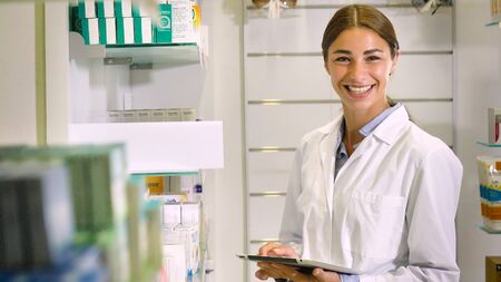 Portrait of a beautiful young girl (woman) pharmacist, consultant, working at a pharmacy, smiling and giving medication. Concept: profession, medecine, medical education.の写真素材