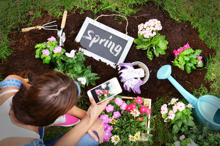 A woman, while using the phone, makes flowers and flowers to give color and decorate her garden, with various tools and a small blackboard with "Spring" writing. Concept of: gardening, appの写真素材