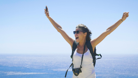 A tourist photographer holds a phone, feels free with a backpack on her back, a wonderfully beautiful view of the blue sea and sky. Concept: travel, vacations, freedom, telephone communication.の写真素材