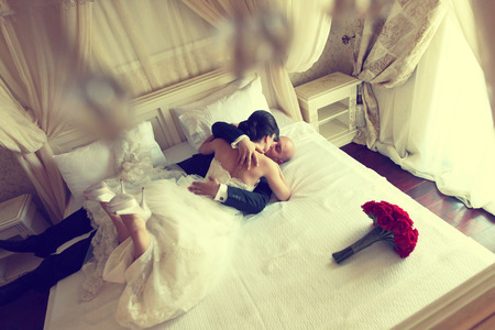 Wedding shot of bride and groom lying in a stylish white bed red flower bouquetの写真素材