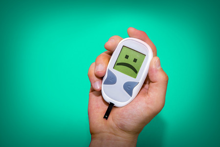 Human hand holding glucometer on turquoise background with sad face on its monitor illustrating bad blood sugar levels. Diabetes conceptの写真素材