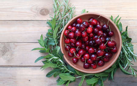 sweet cherry in a ceramic cup with mint and rosemary rim. On a wooden, light brown background. Organic food, place for text.の写真素材