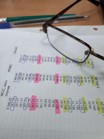 Workplace of an office worker: glasses, business papers with digital data, a pen on a wooden table.