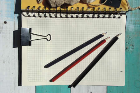 A notebook with white and yellow pages, lying on a blue wooden table. Near pencils, a stone, a shell...