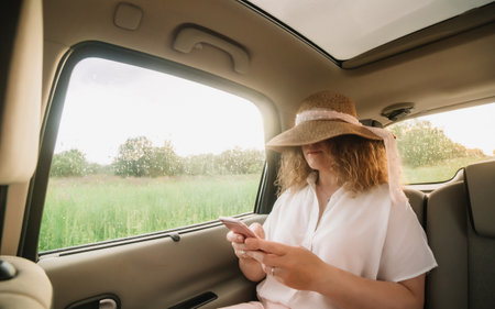 Cheerful positive curly young woman in casual wear with hat sitting in automobile backseat with fastened seatbelt and using phoneの写真素材