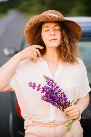Young woman stands in white shirt near car with purple and pink lupins. Beautiful young woman in hat with curly hair with bouquet of lupins. Sunset or sunrise, bright evening light.の写真素材