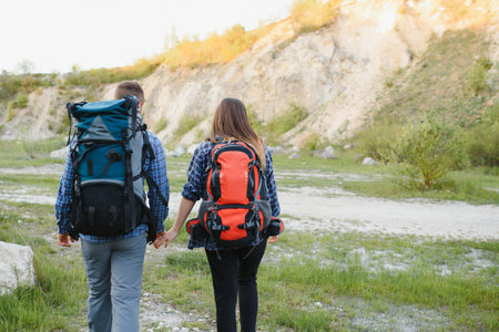 Back view of young pair backpackers with big backpacks holding hands and walking along a road with beautiful mountain landscape on background.の写真素材
