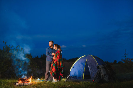 Loving couple hikers enjoying each other, standing by campfire at night under evening sky near trees and tent. Romantic camping near forest in the mountainsの写真素材