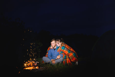 Young couple man and woman travelers sitting near glowing tourist tent, burning campfire, on the top of mountain, enjoying beautiful view of night sky.の写真素材
