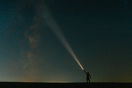 Back view of man with head flashlight standing on green grassy field under beautiful dark blue summer starry sky. Night photography, beauty of nature concept. Wide panorama, copy space background.の写真素材