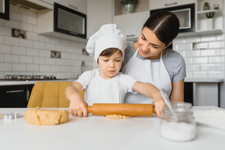 Little boy helping his mother with the baking in the kitchen standing at the counter alongside her...