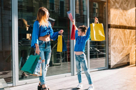 Beautiful mom and her cute little daughter are holding shopping bags, looking at camera and smiling while standing outdoors. shopping concept.の写真素材