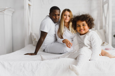 Portrait of happy multiracial young family lying on cozy white bed at home, smiling international mom and dad relaxing with little biracial girl child posing for picture in bedroomの写真素材