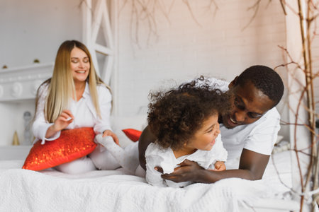 Portrait of happy multiracial young family lying on cozy white bed at home, smiling international mom and dad relaxing with little biracial girl child posing for picture in bedroomの写真素材