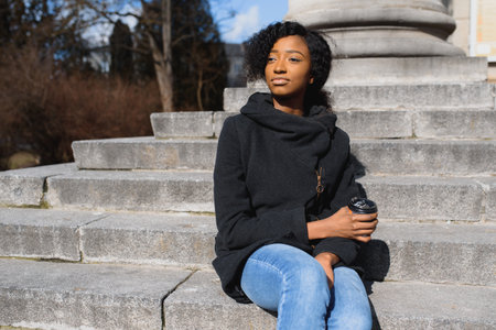 Pensive african woman wearing glasses drinking take away coffee sitting on the street stairsの写真素材