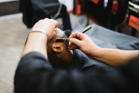 Side view of serious man with stylish modern haircut looking forward in barber shop. Hand of barber keeping straight razor and cutting trendy stripes on head of client.の写真素材