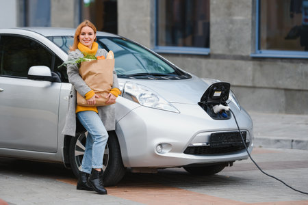 Woman with a mobile phone near recharging electric car. Vehicle charging at public charging station outdoors. car sharing conceptの写真素材