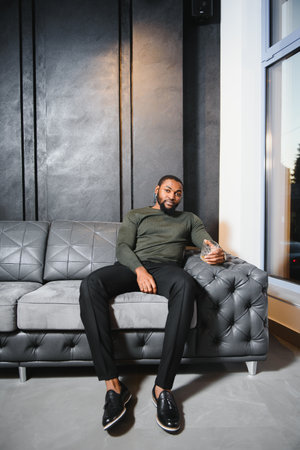 Handsome young african american man in elegant suit with glass of whiskey relaxing on sofa