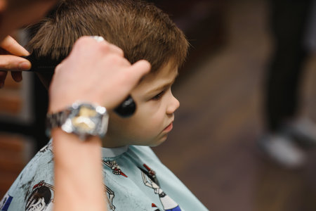 A little cute boy sits in a hairdresser's at the stylist's, a schoolchild is getting hair cut in a beauty salon, a child at a barbershop's, a short men's haircut.の写真素材