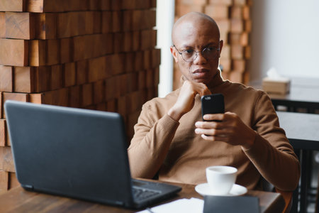 African businessman talking on phone sitting at cafe table, busy entrepreneur working distantly in coffee house with laptop papers speaking on mobile, black man making call having lunch in cafeteriaの写真素材