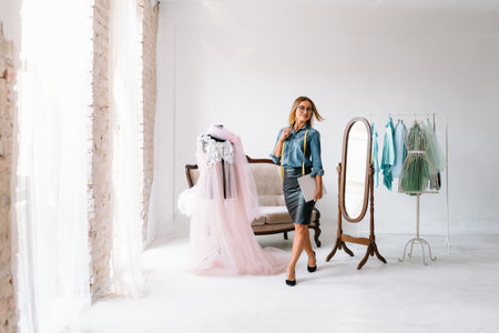 Smiling fashion designer looking at camera at workplace, dressmaker, needlewoman or tailor shop owner sitting at desk with color swatches and embroidery design sketches on the wall, portrait.の写真素材