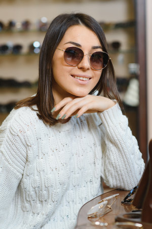 Portrait of a young woman shopping, standing in store and trying sunglasses near a mirror.の写真素材