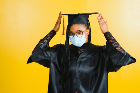 Portrait of a young african student in a graduate cap protective mask, on a yellow background, Graduation 2021の写真素材