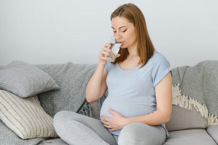 Pregnant woman with glass of water sitting on sofa. Young expectant blonde rest at home, copy space....