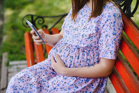 Pregnat woman sits on bench in the park and using phone. Happy female have rest outdoor during the...