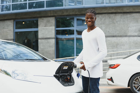 african man holding charge cable in on hand standing near luxury electric car.
