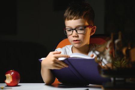 concentrated schoolboy reading book at table with books, plant, lamp, color pencils, apple, and...