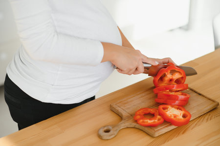 pregnancy, cooking food, healthy lifestyle, people and expectation concept - happy pregnant woman with knife chopping cucumber and preparing vegetable salad at homeの写真素材