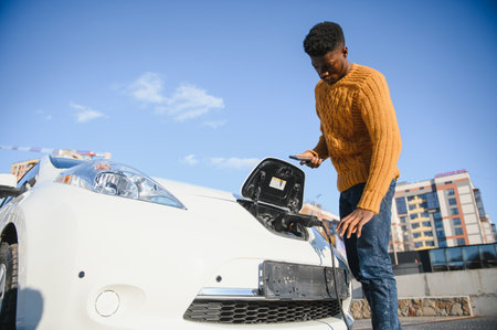 Close up of african american man connecting charging cable to electric car. Young male standing near...