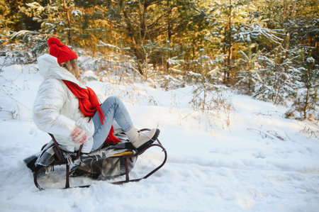 Beautiful young woman wearing a warm winter clothes, sitting on a sleigh and sliding down the hill,...