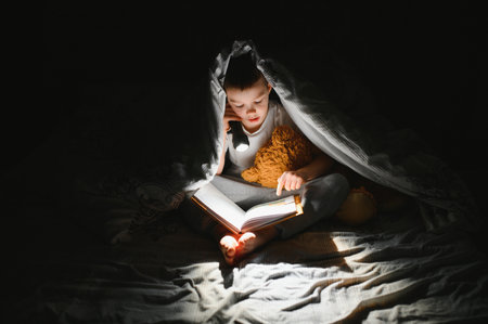 Reading book and using flashlight. Young boy in casual clothes lying down near tent at evening time.の写真素材