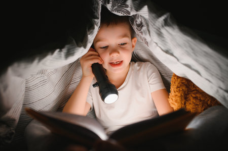 Portrait of cute little boy reading in bed with flashlight in dark room, enjoying fairytales.の写真素材