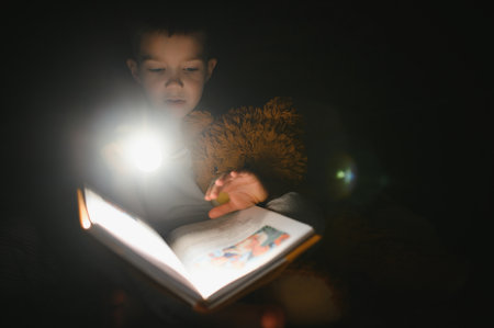 Child reading book in bed. Kids read at night. Little boy with fairy tale books in the bedroom. Education for young children. Bedtime story in the evening. Cute kid under blanket in dark room with lamp.の写真素材