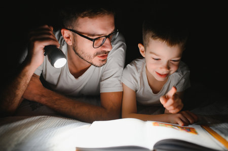 Father and son with flashlight reading book under blanket at homeの写真素材